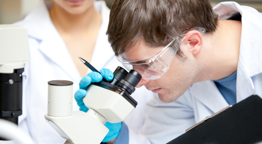 Caucasian male scientist holding pen and clipboard looking through a microscope in his laboratory size m